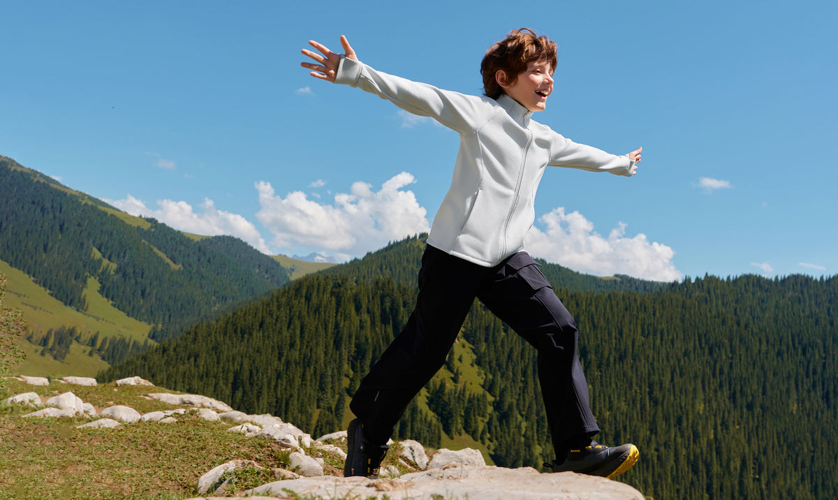 Boy in a light fleece jacket and black pants leaping on a mountain ridge